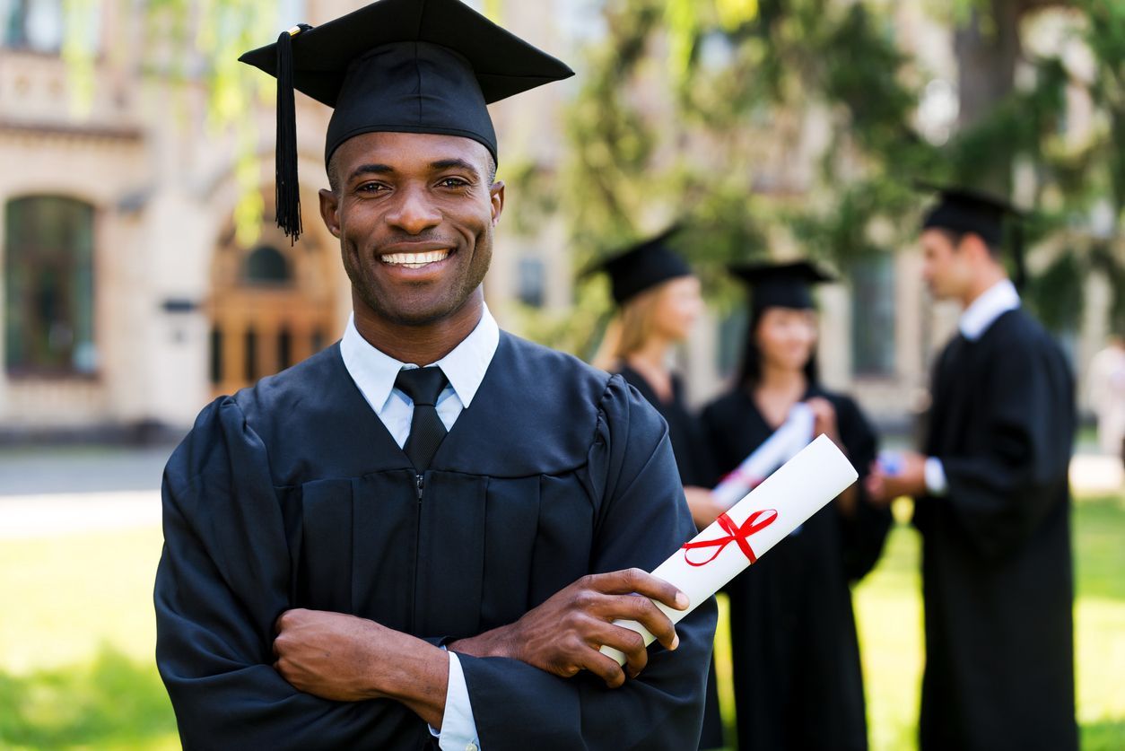 Smiling Black graduate in cap and gown holding diploma, other graduates and a university building in the background.