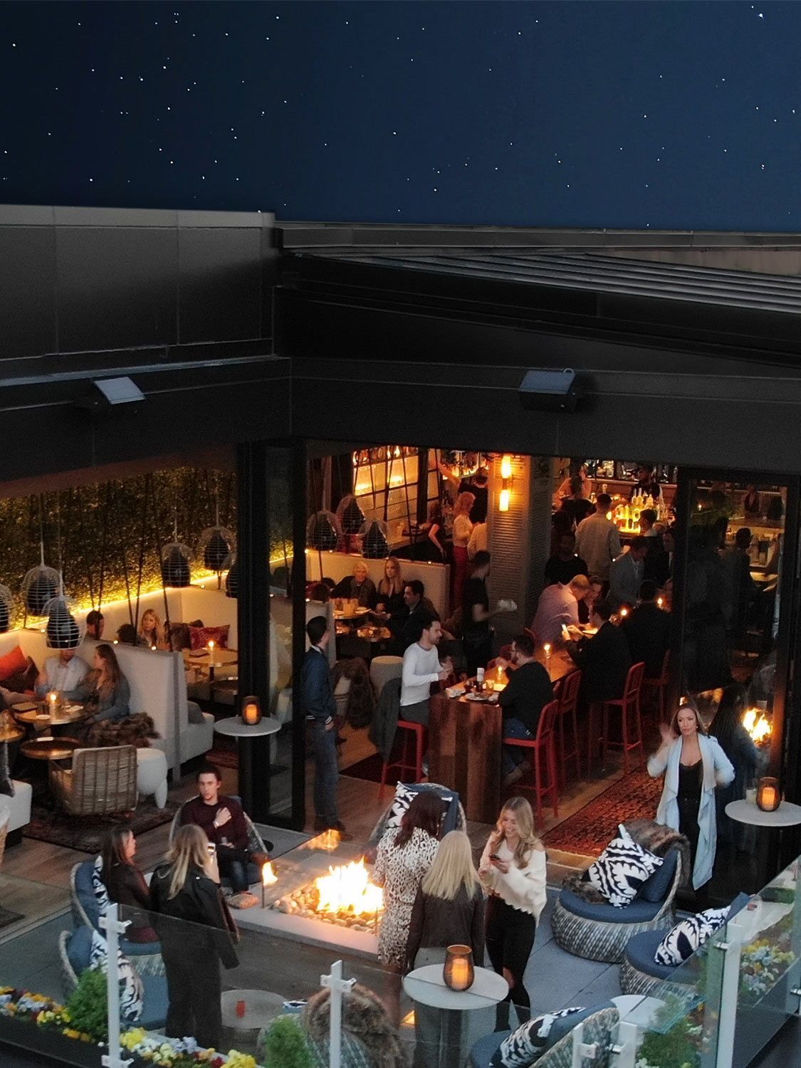 Night scene: Rooftop restaurant with outdoor fire pit and bar. People dining and socializing under starry sky.
