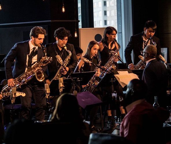 A jazz band, playing saxophones, performs in a dimly lit venue for an audience. Musicians wear dark suits.