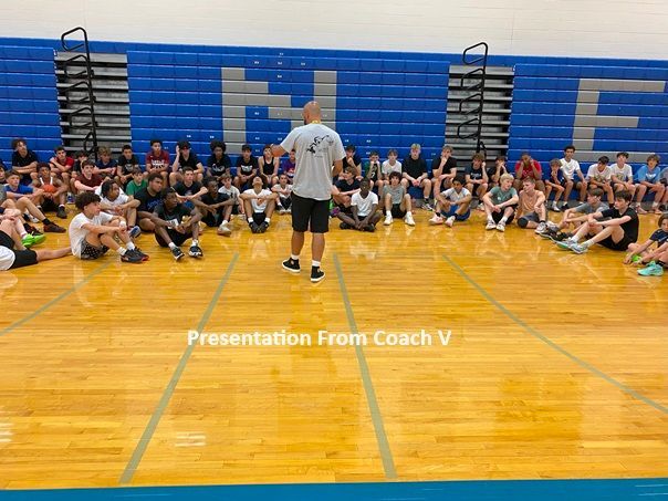 A man is standing in front of a group of people sitting on the floor in a gym.