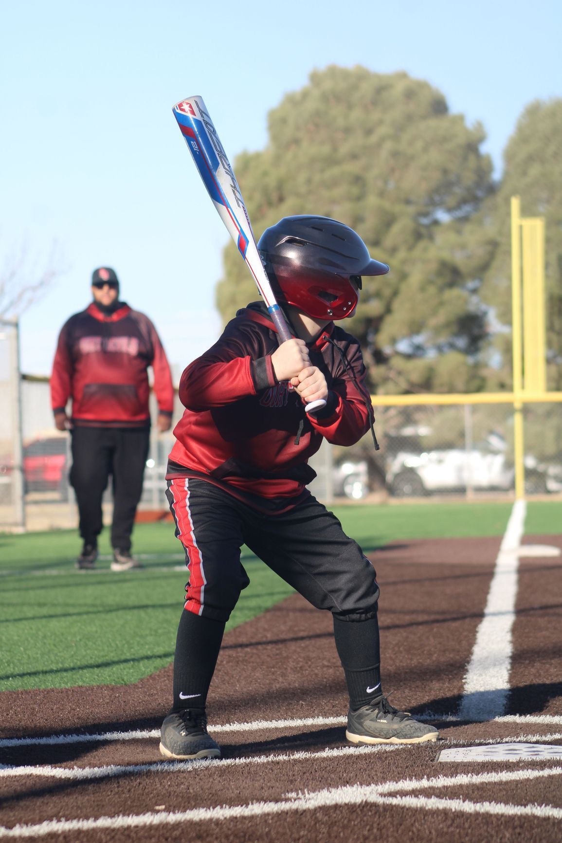 Young baseball player at bat, wearing a helmet and red/black uniform. A coach stands in the background on a baseball field.