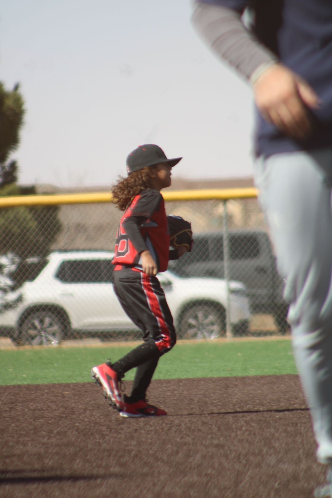 A young baseball player with long hair runs on a dirt infield, wearing a black cap, red and black uniform, and holding a glove.