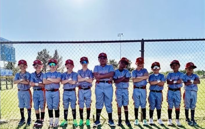 Youth baseball team posing in uniform, standing in front of a fence. They are wearing red and blue uniforms with baseball caps, some with sunglasses.