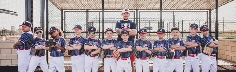 A youth baseball team poses for a team photo. The players and coach stand with arms crossed in front of a chain-link fence.