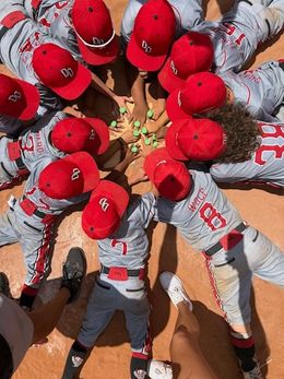 A youth baseball team in gray uniforms and red hats gathers in a circle, hands in the center, on a dirt field.