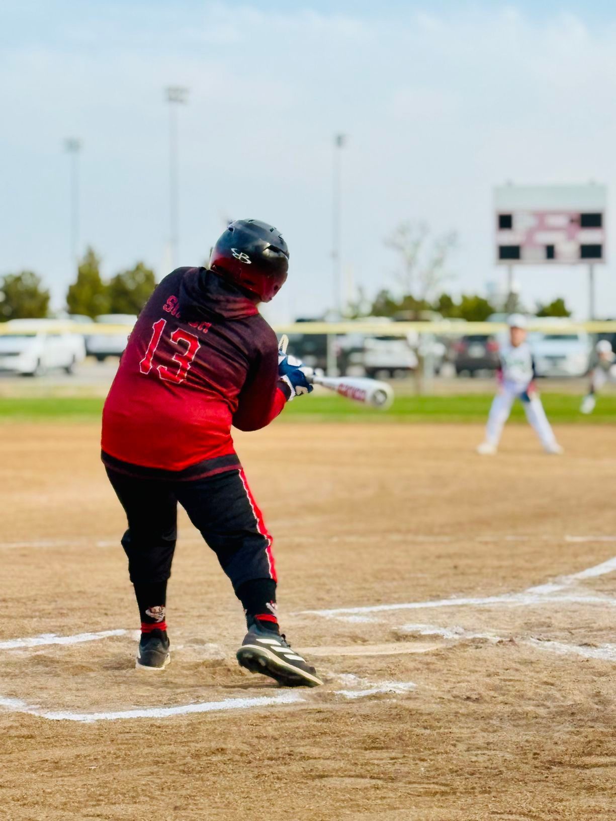 Softball player in a red jersey with the number 13 swings a bat at the plate. The batter is facing the pitcher in a ballpark.