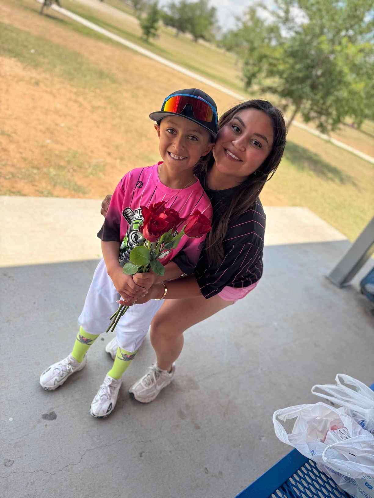 A smiling woman embraces a boy holding red roses at a park. The boy wears a baseball cap and jersey.