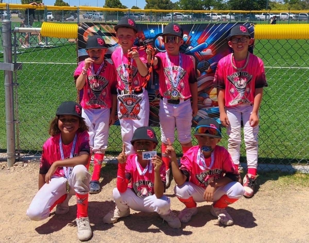 Youth baseball team in pink uniforms and hats poses on a baseball field. They're smiling, some holding medals, in front of a fence.