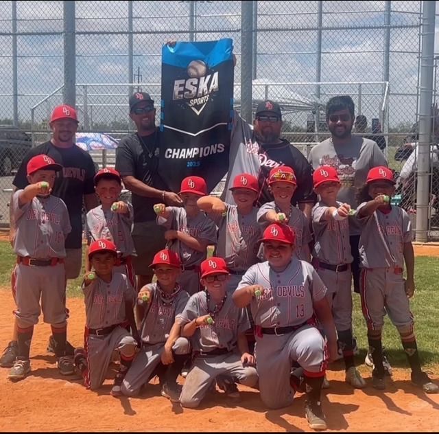 Youth baseball team in red uniforms celebrates a championship victory, posed on a baseball field with coaches and trophy banner.