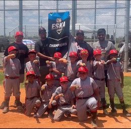 Youth baseball team, in red and gray uniforms, celebrating a championship win on a baseball field, holding a banner that says 