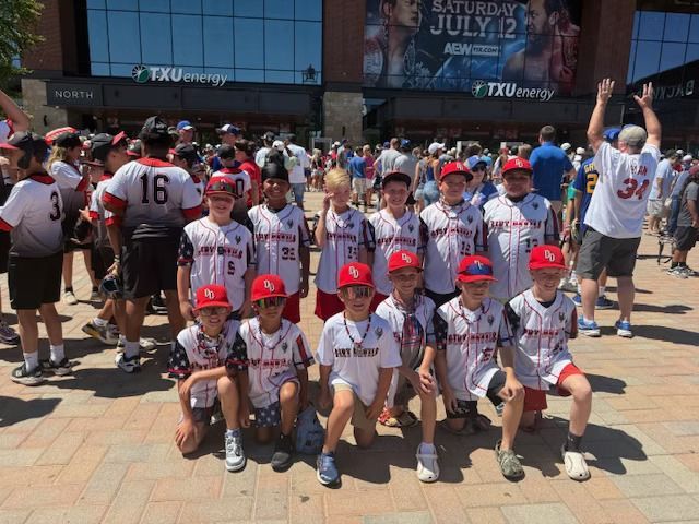 Youth baseball team in matching jerseys and red caps pose in front of a stadium on a sunny day.