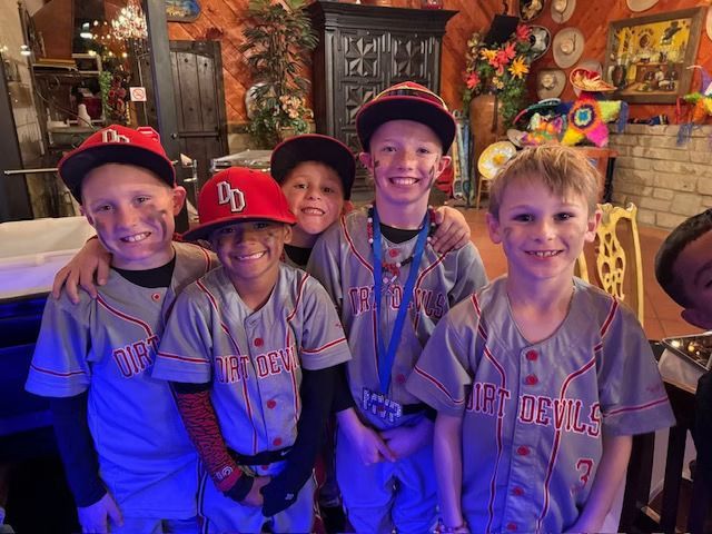 A group of young baseball players in gray and red Dirt Devils uniforms smile for a photo in a restaurant. They have smudged dirt on their faces.