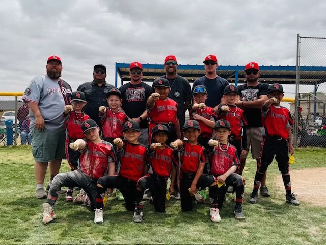 Youth baseball team in red and black uniforms poses for a photo on a baseball field. Coaches stand in the back.