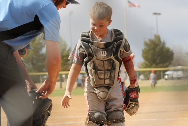 Young baseball catcher, blood on arm, walks off field looking down, coach nearby.
