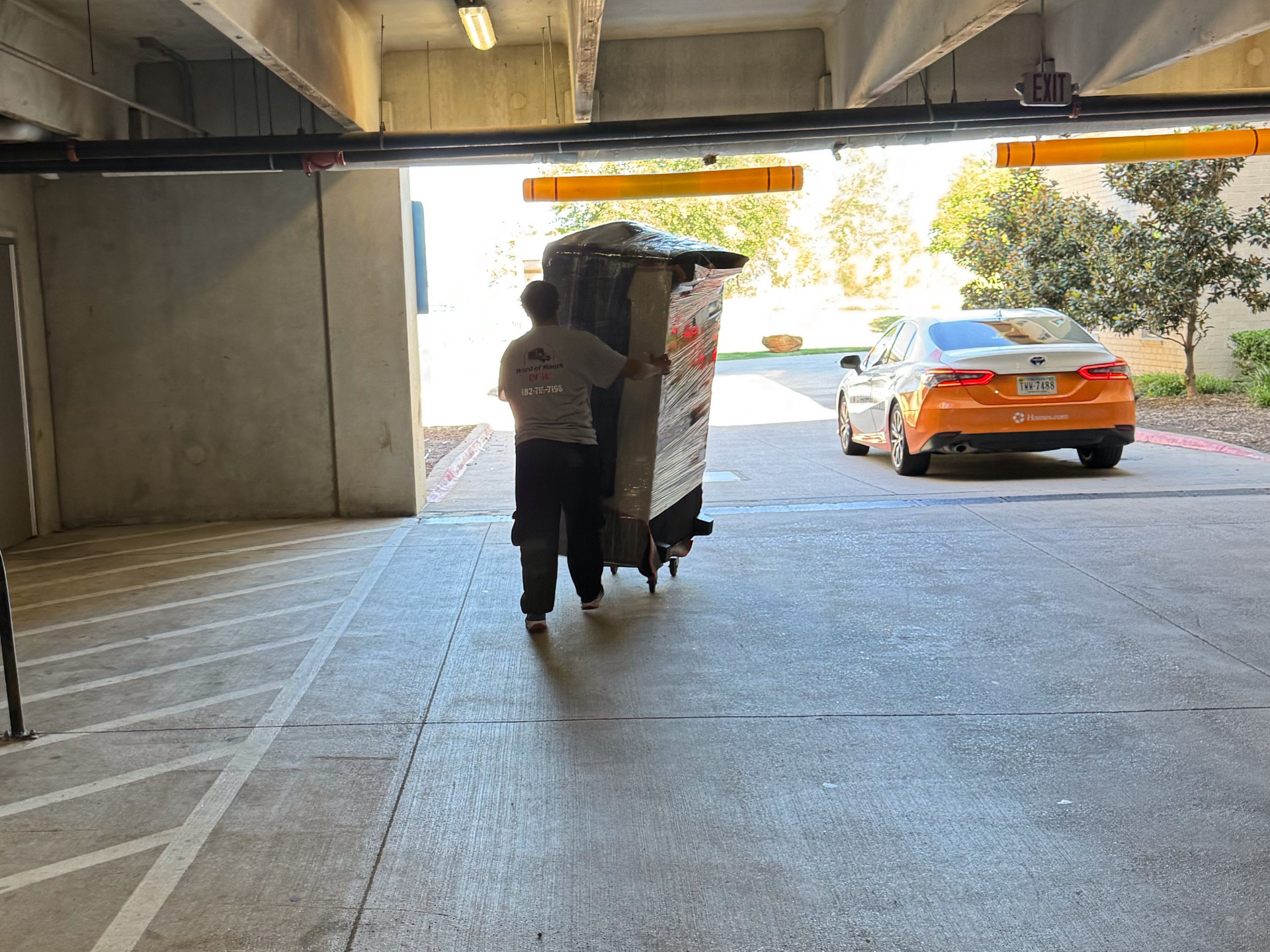 A person pulls a wrapped object on a cart out of a parking garage, orange car parked nearby.