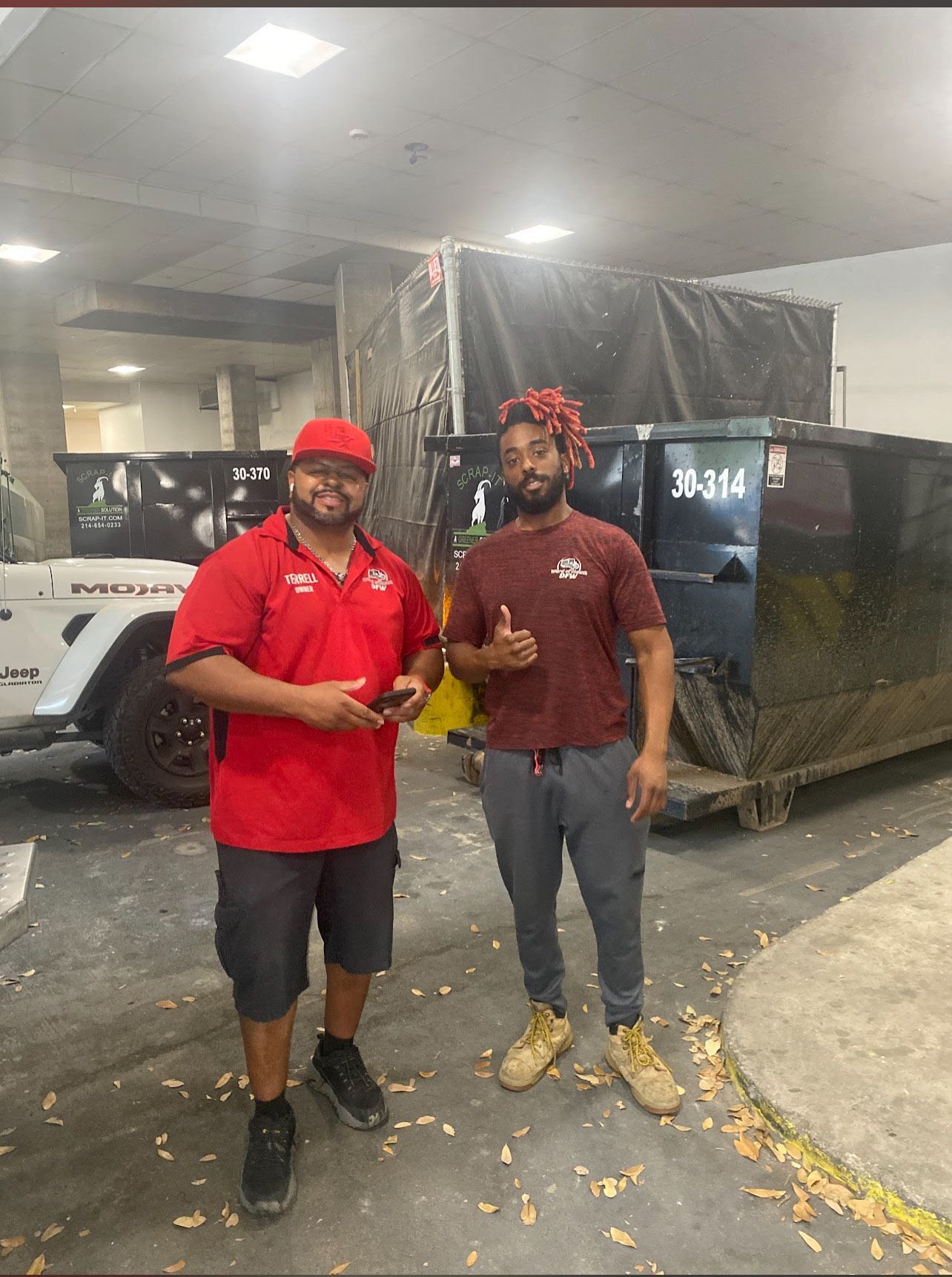 Two men standing near dumpsters. One in red gives a thumbs up. Concrete setting.