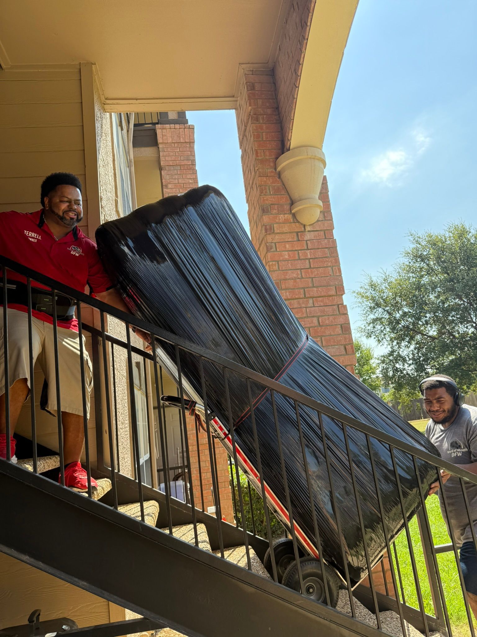 Two men carrying a large, black-wrapped item up exterior stairs; sunny, brick building.