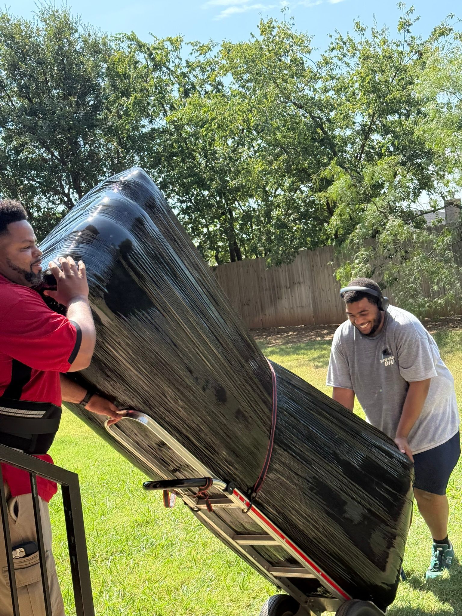 Two men moving a large, wrapped object on a hand truck in a grassy yard. Sunny day.
