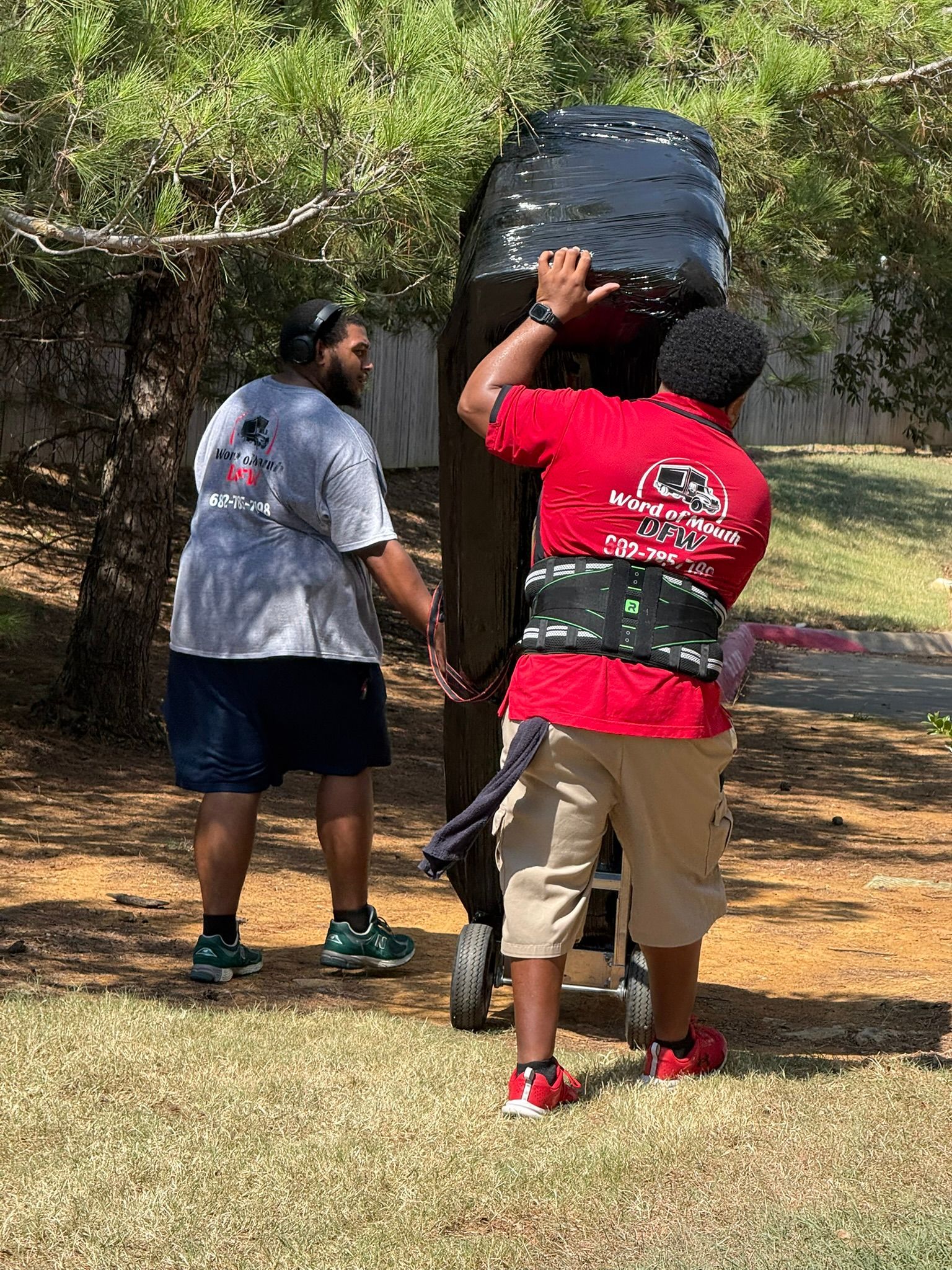 Two men moving a tall black object on a wheeled cart outdoors. One man is wearing a red shirt.