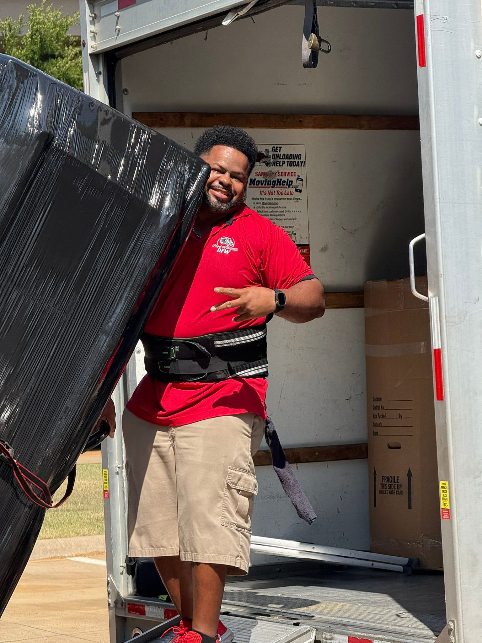 Man in red shirt and cargo shorts, wearing back support, smiles while loading a black, wrapped item into a truck.