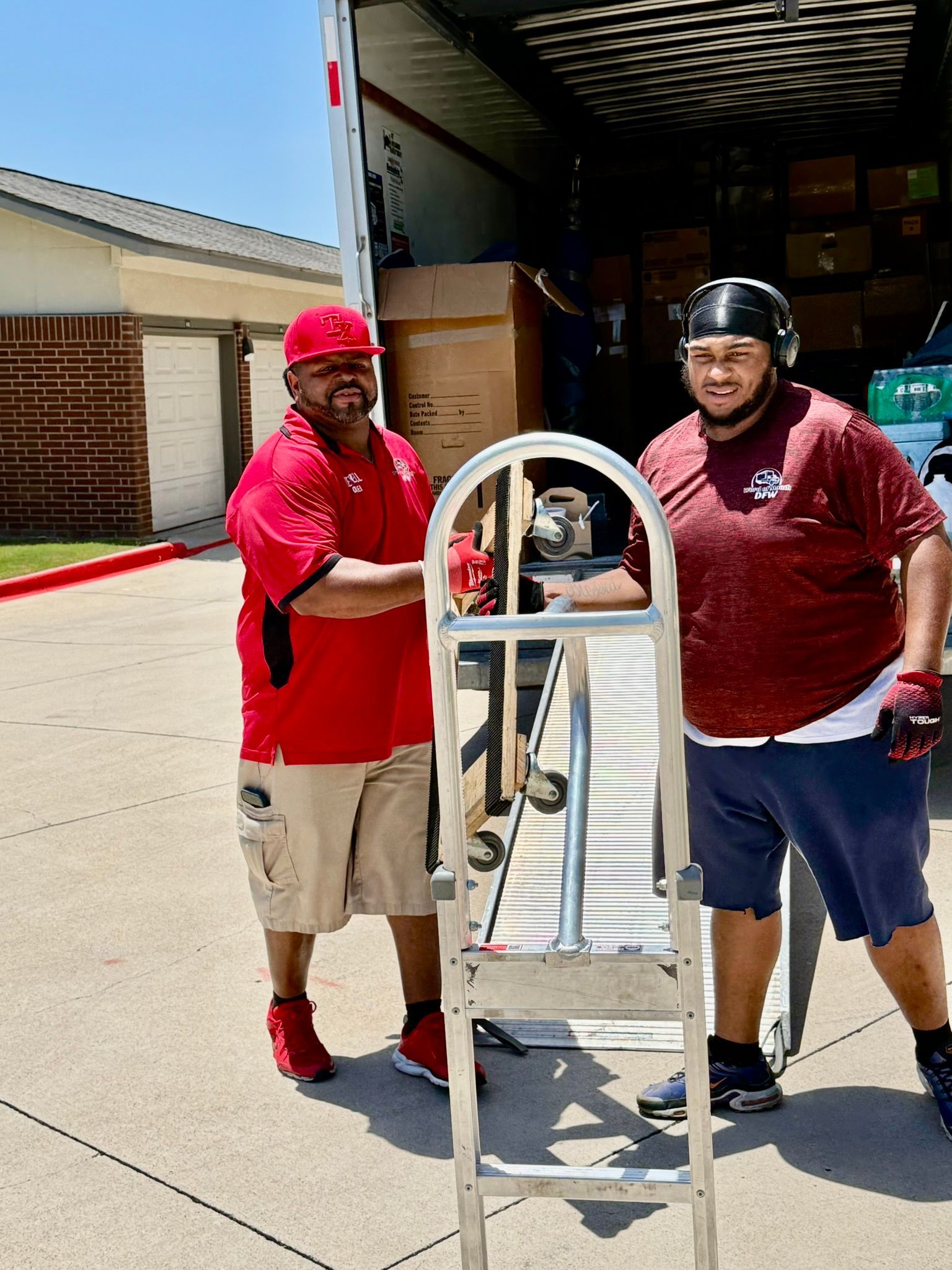 Two movers stand near a truck. One in red leans on a hand truck, the other in red has his arms crossed.