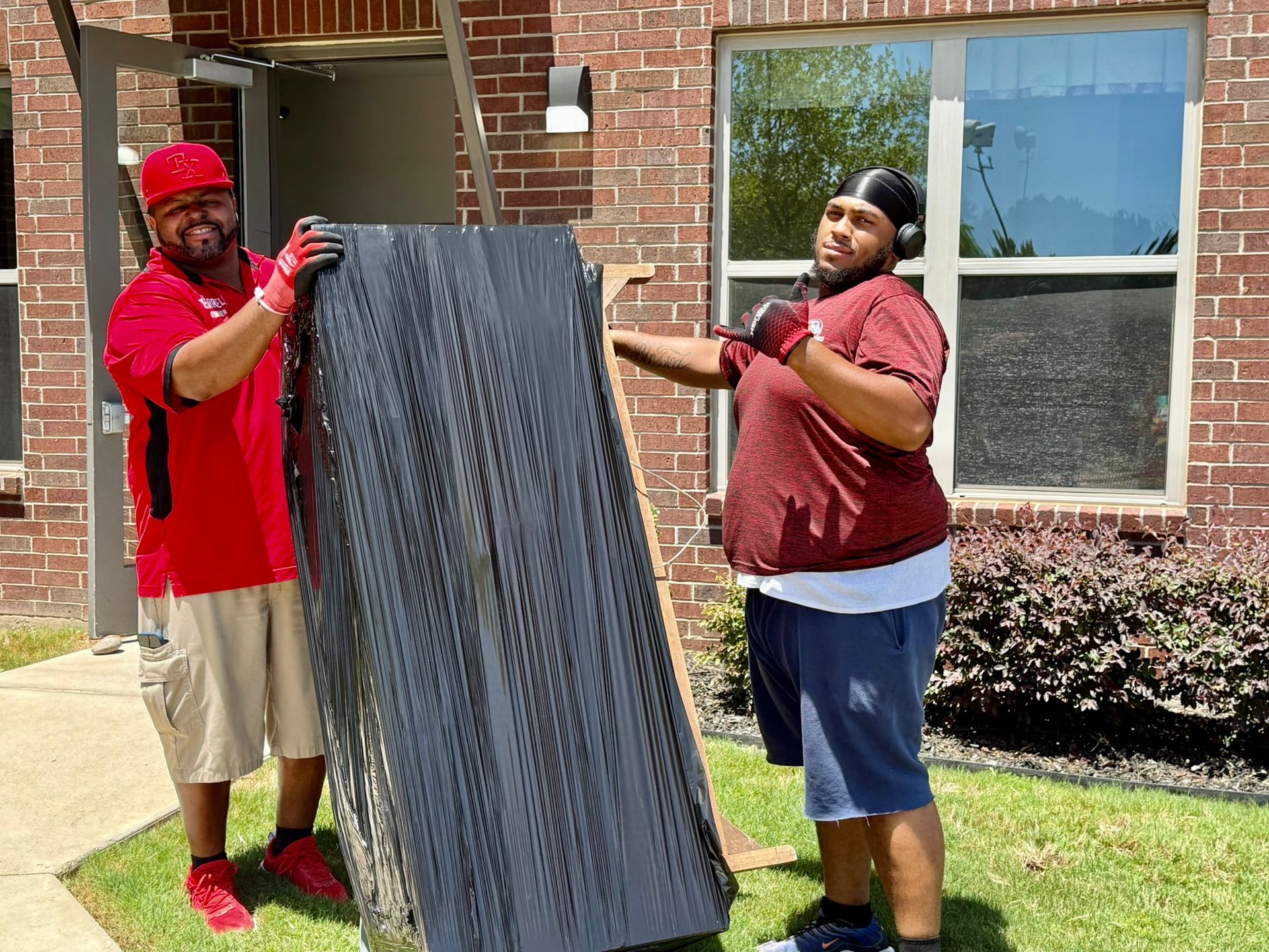 Two men carrying a large, dark panel outside a brick building. One gives a thumbs-up, both wearing gloves.