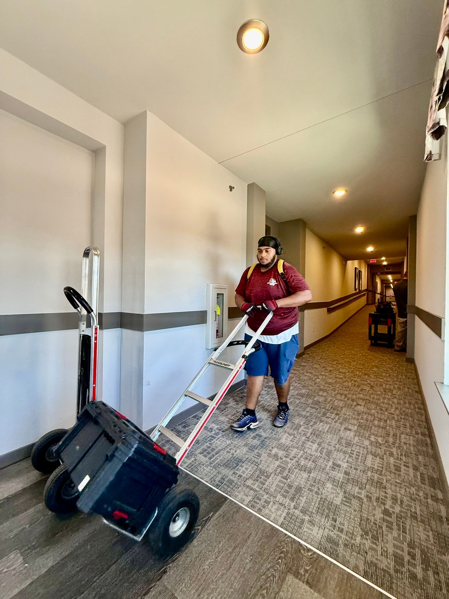 Person in maroon shirt and blue shorts pushing a cart down a hallway with patterned carpet.