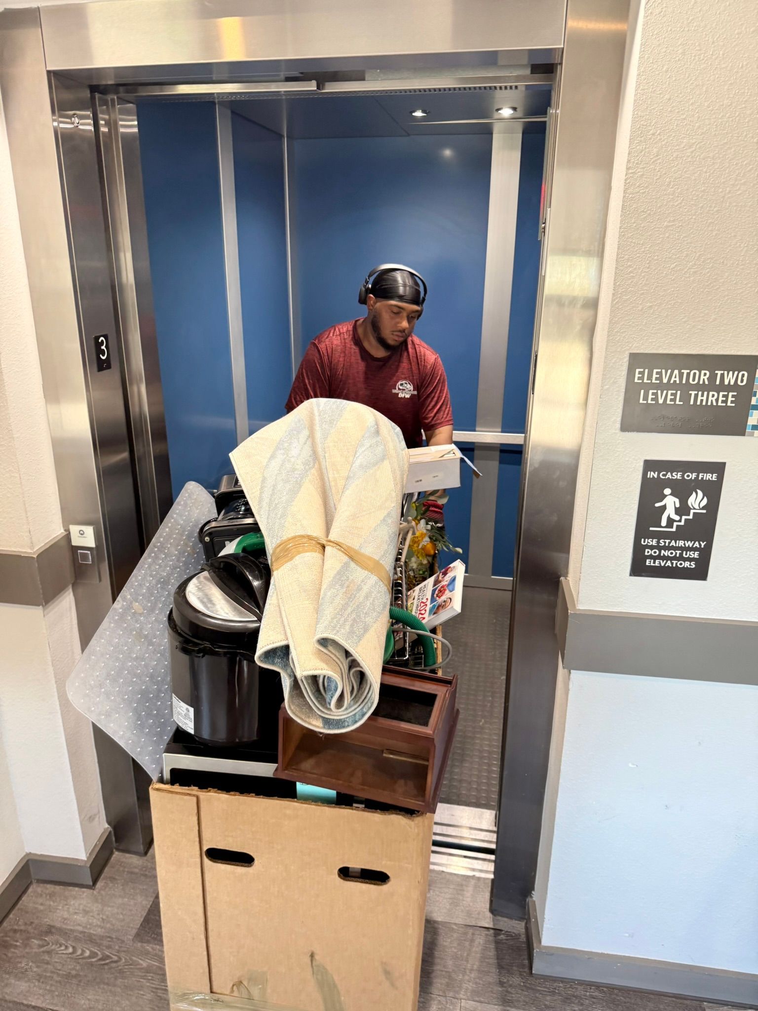 Man in maroon shirt in elevator, surrounded by boxes and household items.