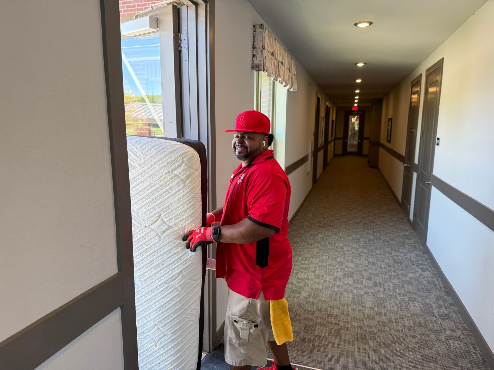 Man in red shirt and hat carrying a mattress through a hallway.