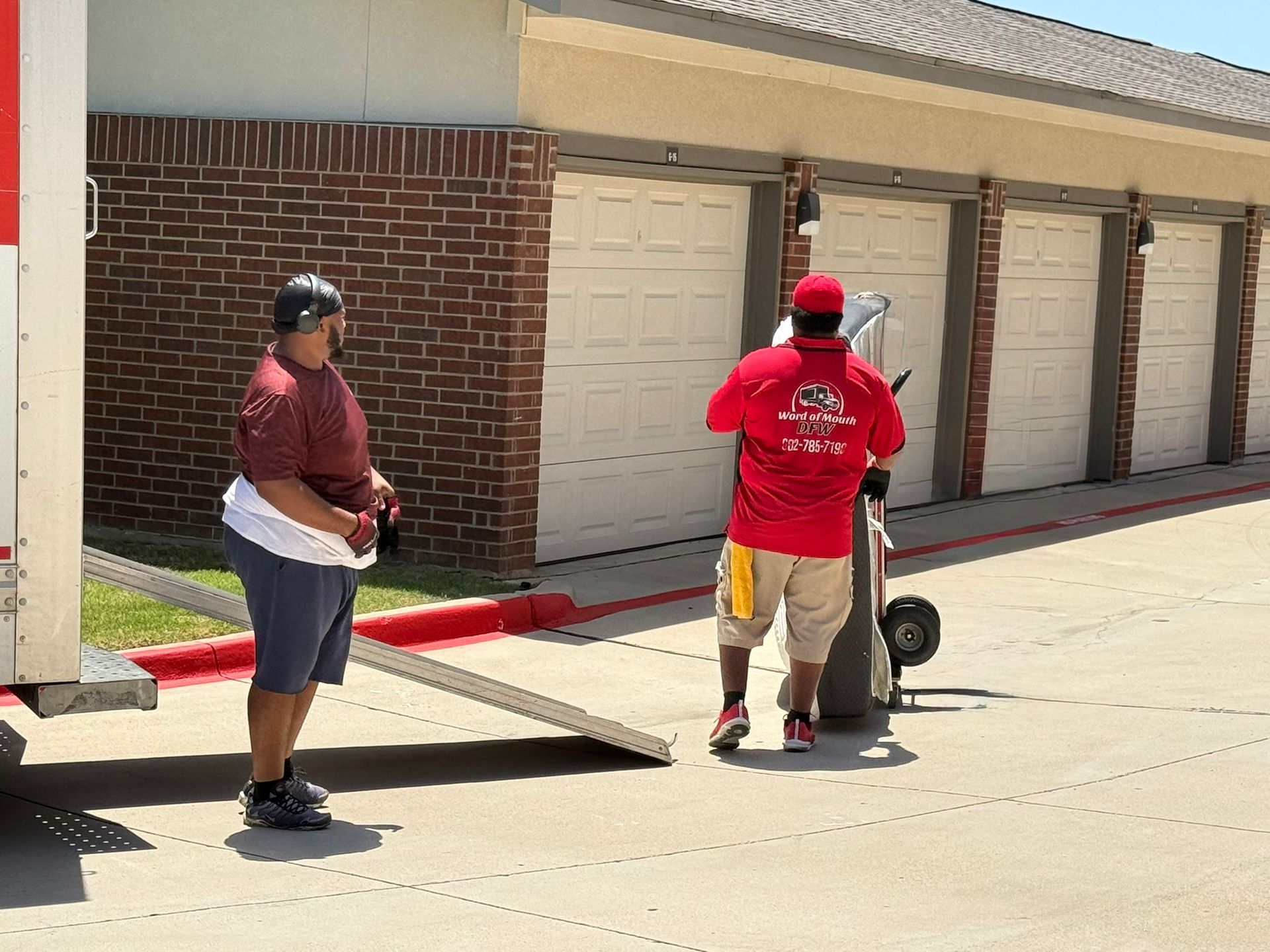 Two movers unloading a truck at storage units. One uses a dolly. Brick building, sunny day.