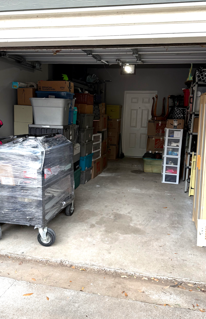 Cluttered garage: boxes, containers, and a cart fill the space. A white door is visible at the back.
