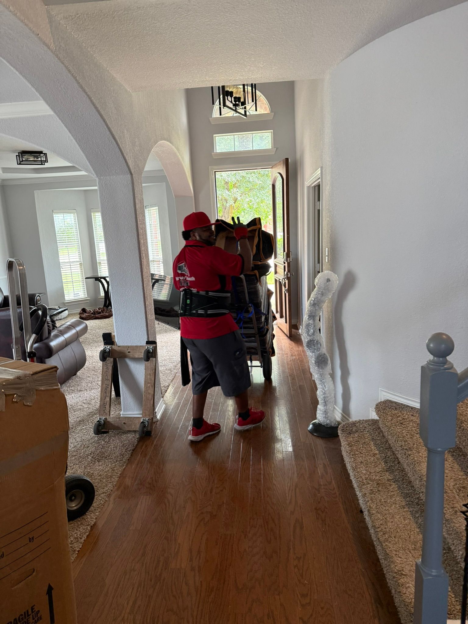 A mover wearing a red shirt, loading belongings on a dolly through a house doorway.