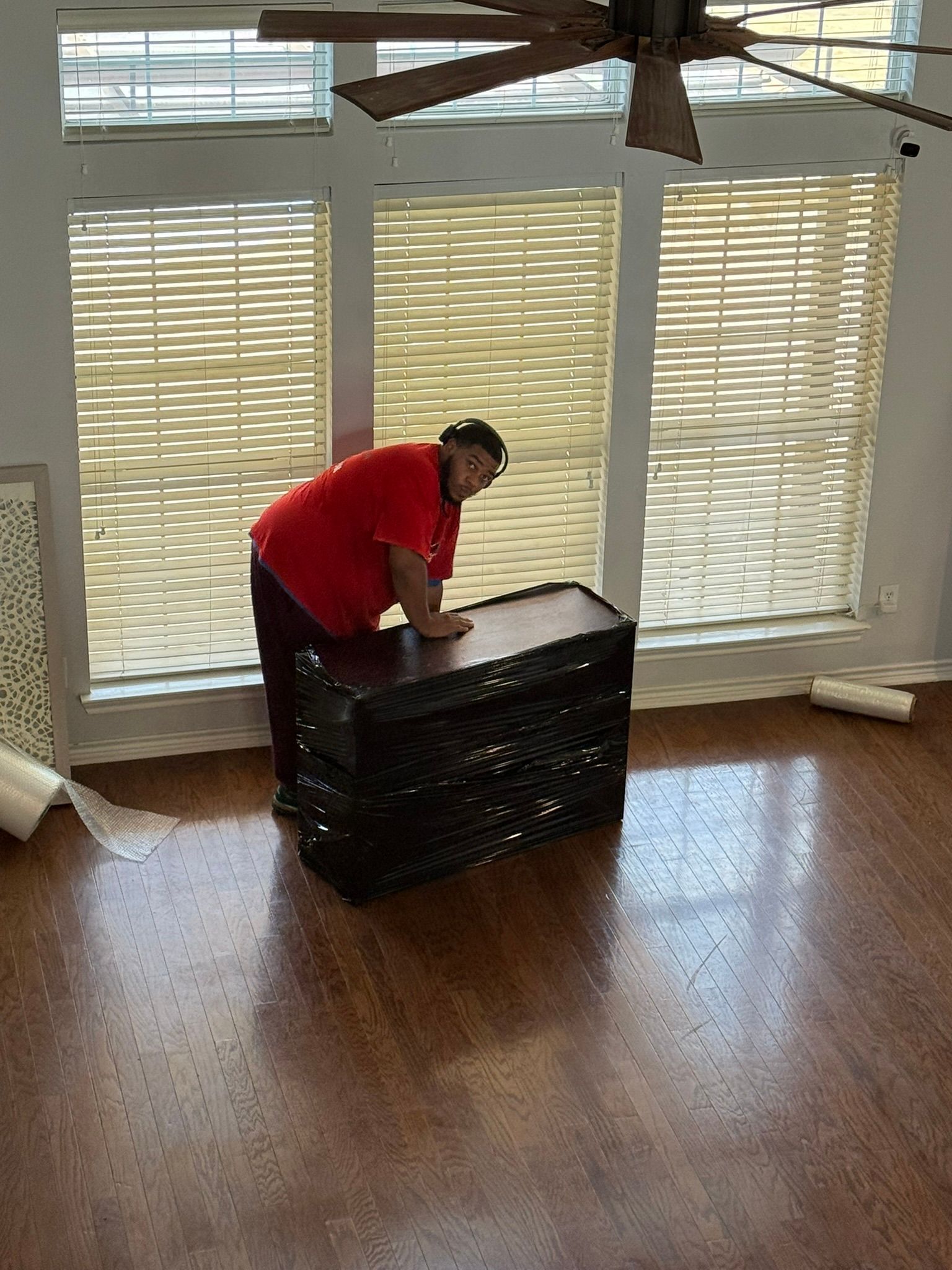 Woman in red shirt moving a wrapped dark chest in a room with large windows.