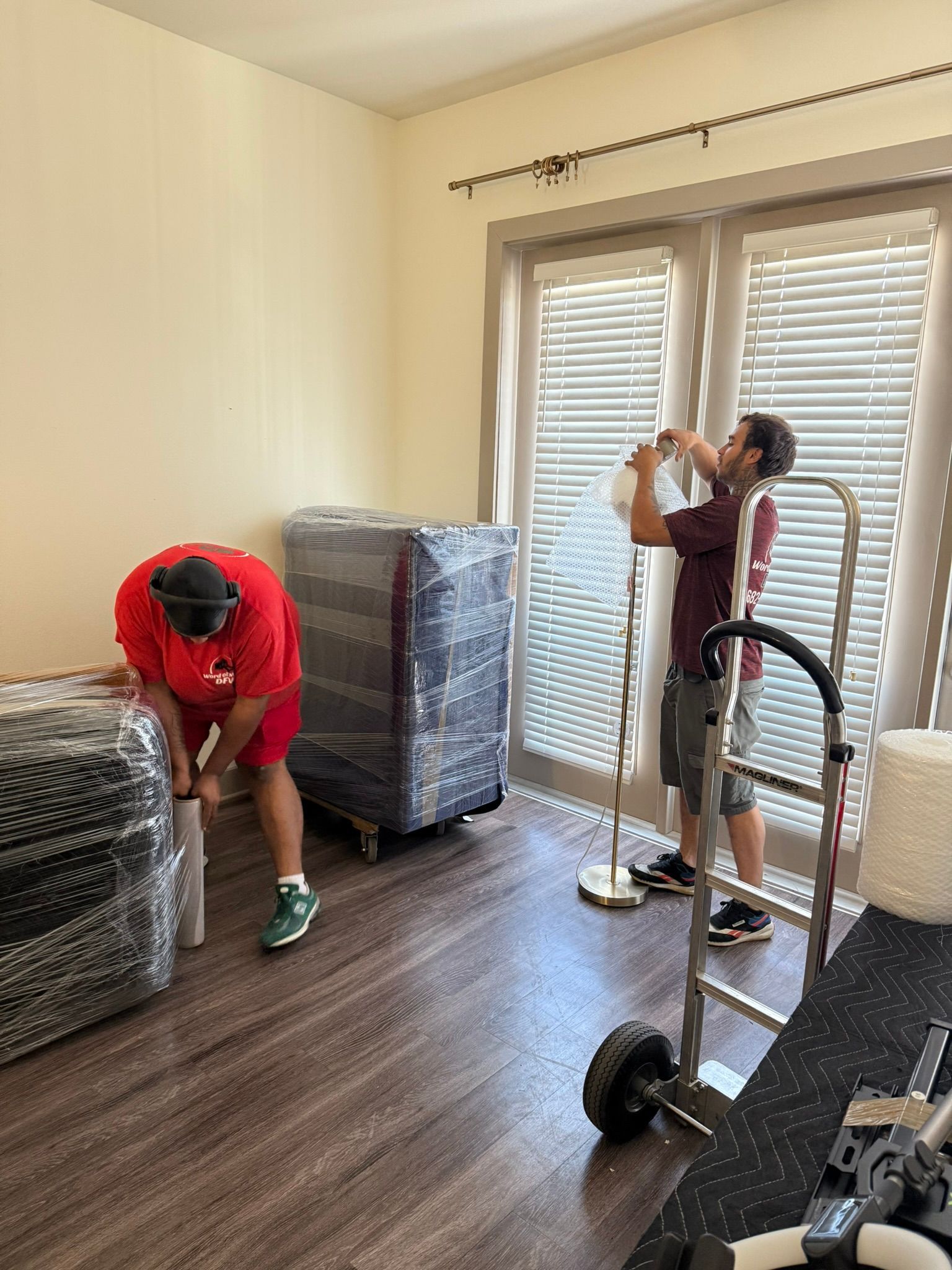 Two movers wrapping furniture inside a room with blinds. One man tapes a lamp, another handles a chair.
