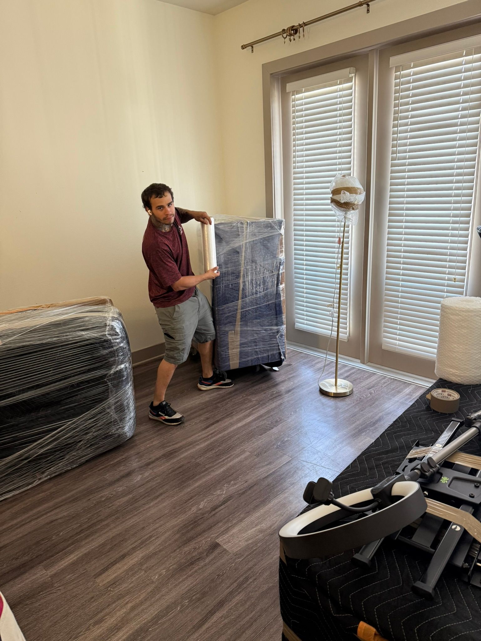 Man moving a wrapped furniture piece on a dolly indoors. He's struggling, beige walls and window in background.