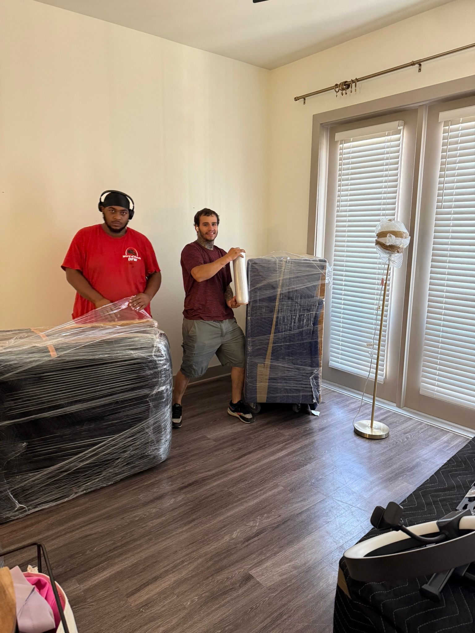 Two movers transport wrapped furniture into a room. One man points; room has dark floors, a lamp, and a door.