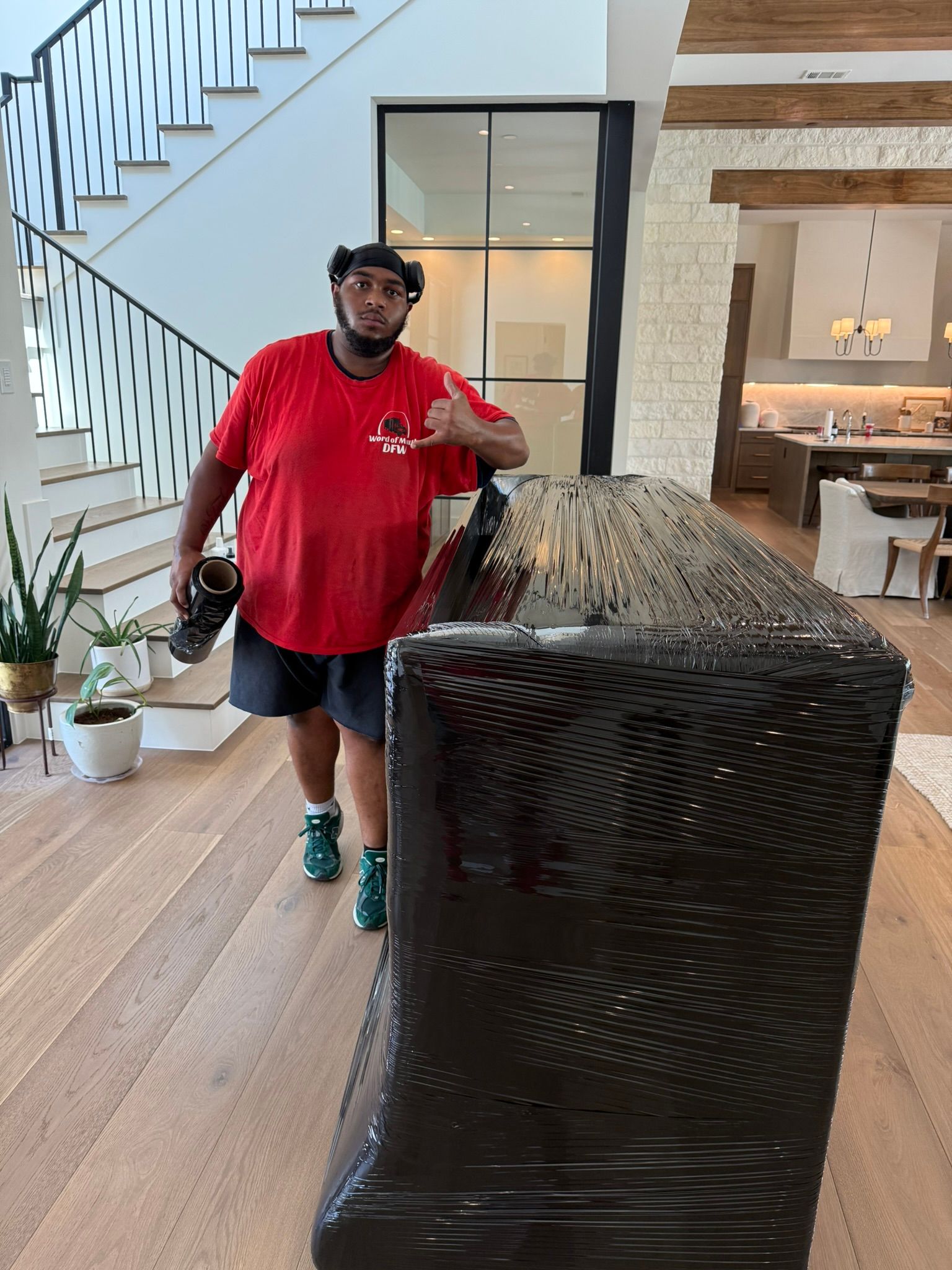 A man in red shirt and shorts moves furniture wrapped in black plastic inside a bright house.