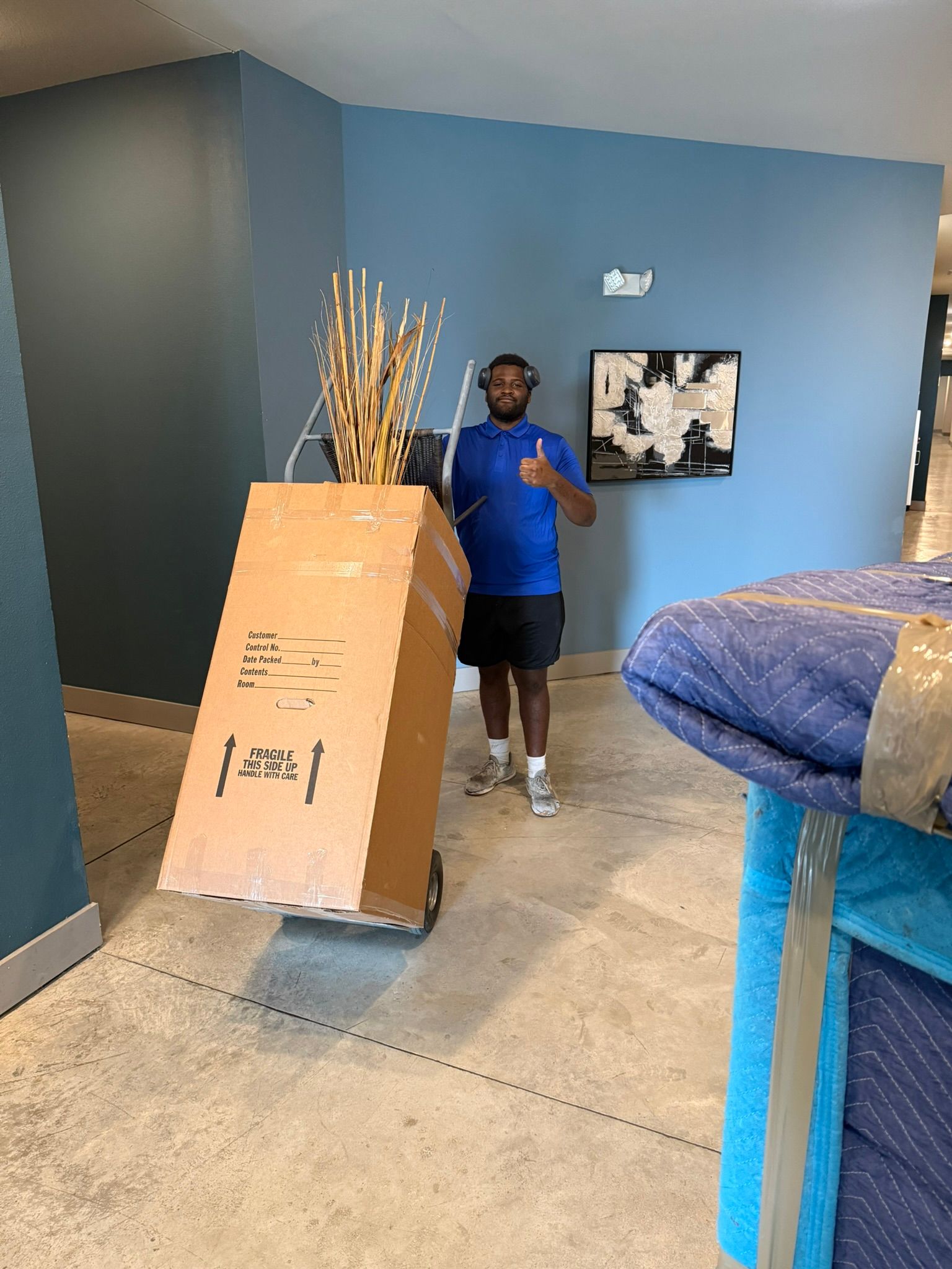 Man pushing a large cardboard box on a dolly. He gives a thumbs up. Blue wall and hallway.