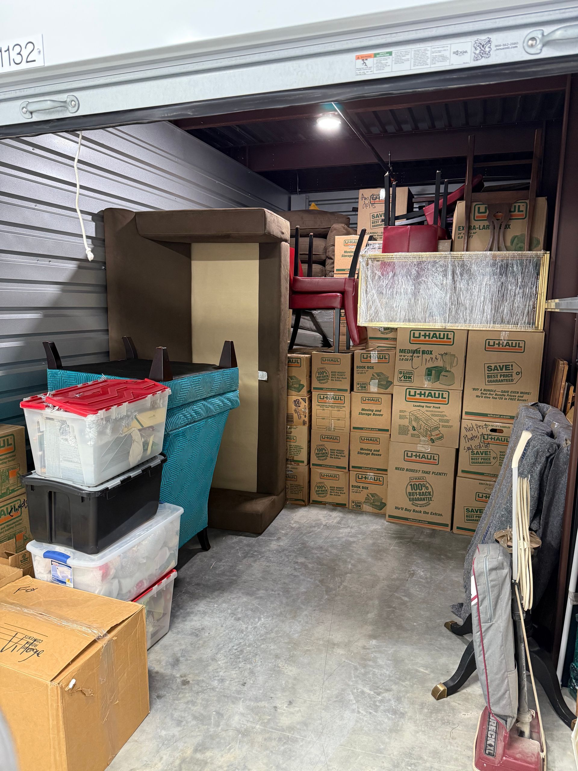 A storage unit filled with packed boxes, furniture, and containers. The door is open.