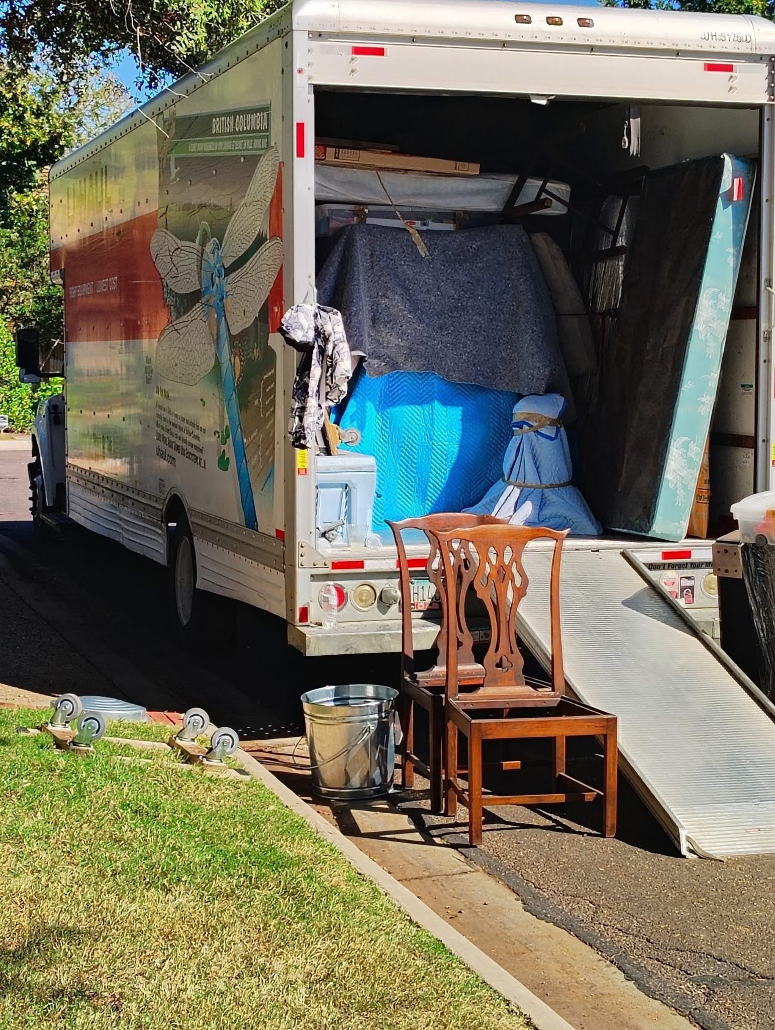 Moving truck loading, open ramp. Chairs and other items outside; truck interior packed with furniture.