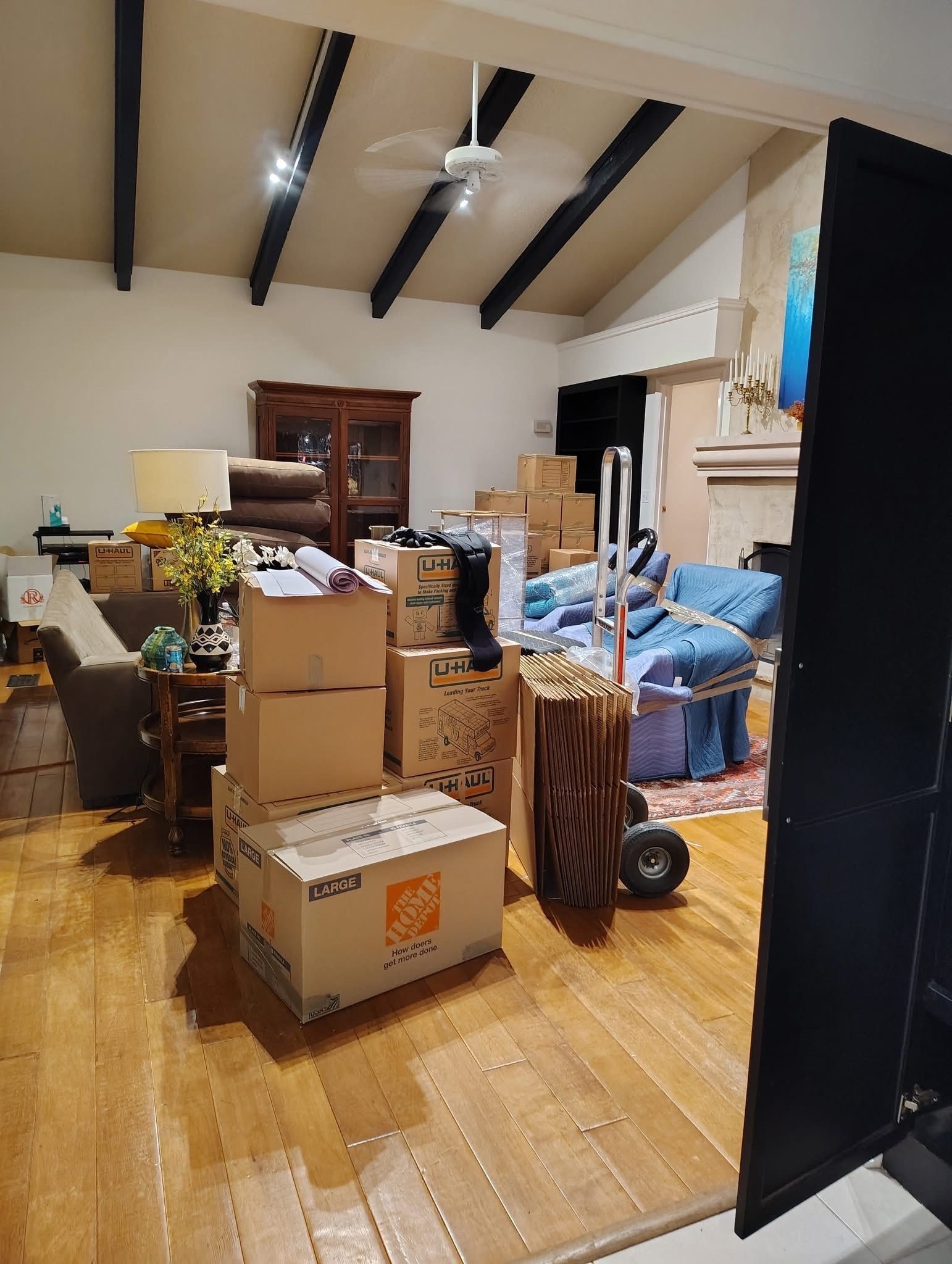 Living room filled with moving boxes and furniture, with a hand truck. Wooden floor and beamed ceiling.