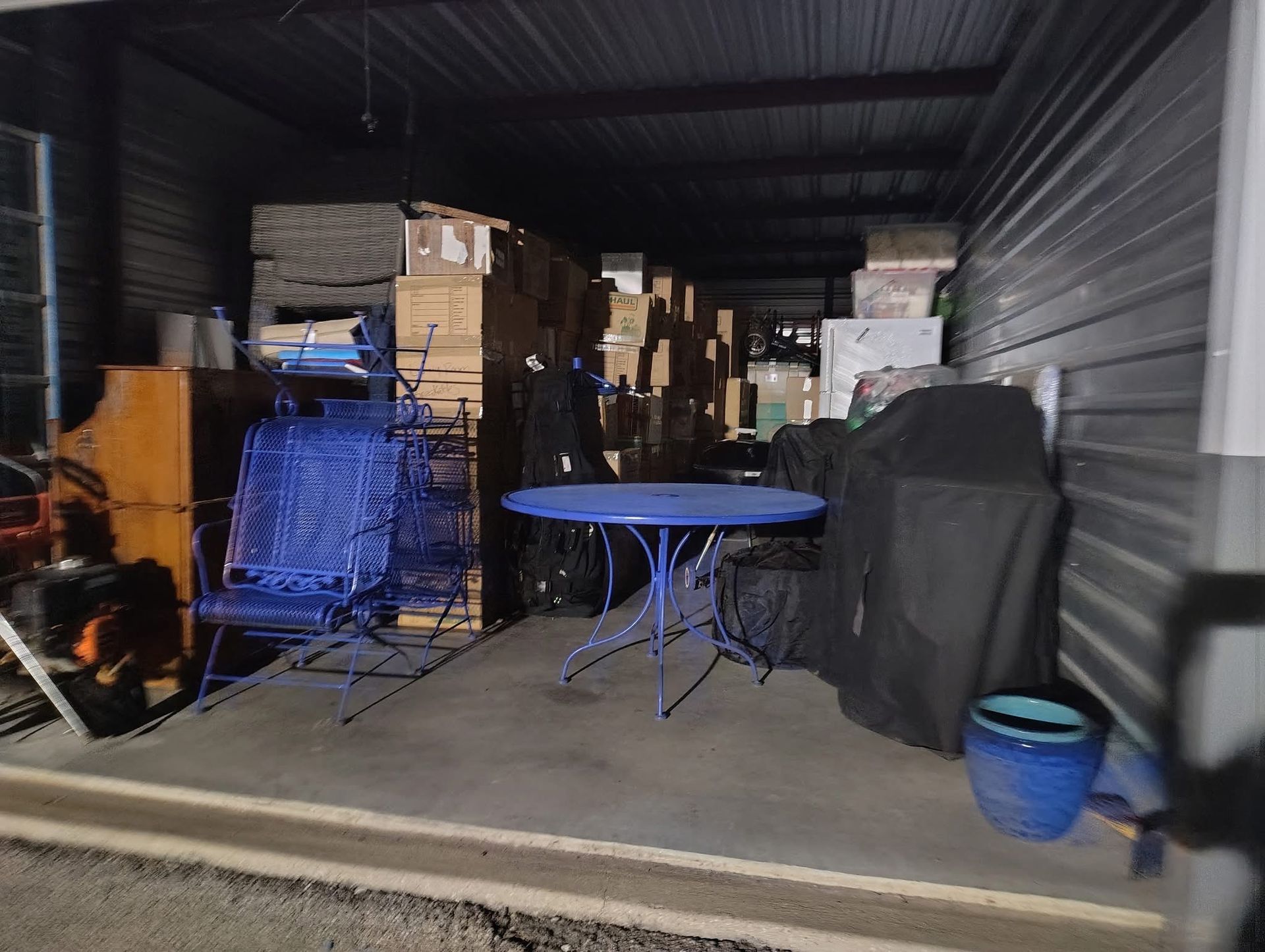 Storage unit with blue table and chairs, various boxes and items. Dark, dimly lit.
