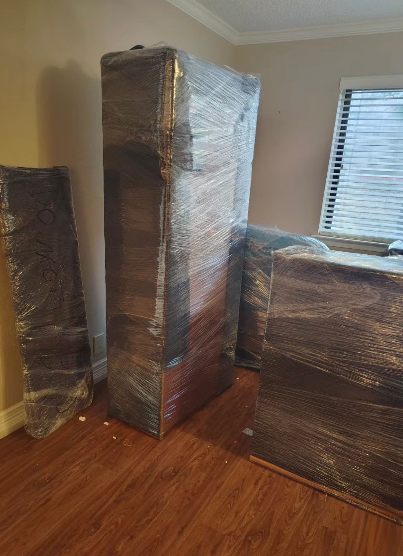 Furniture wrapped in plastic, standing in a room with wood flooring, near a window with blinds.