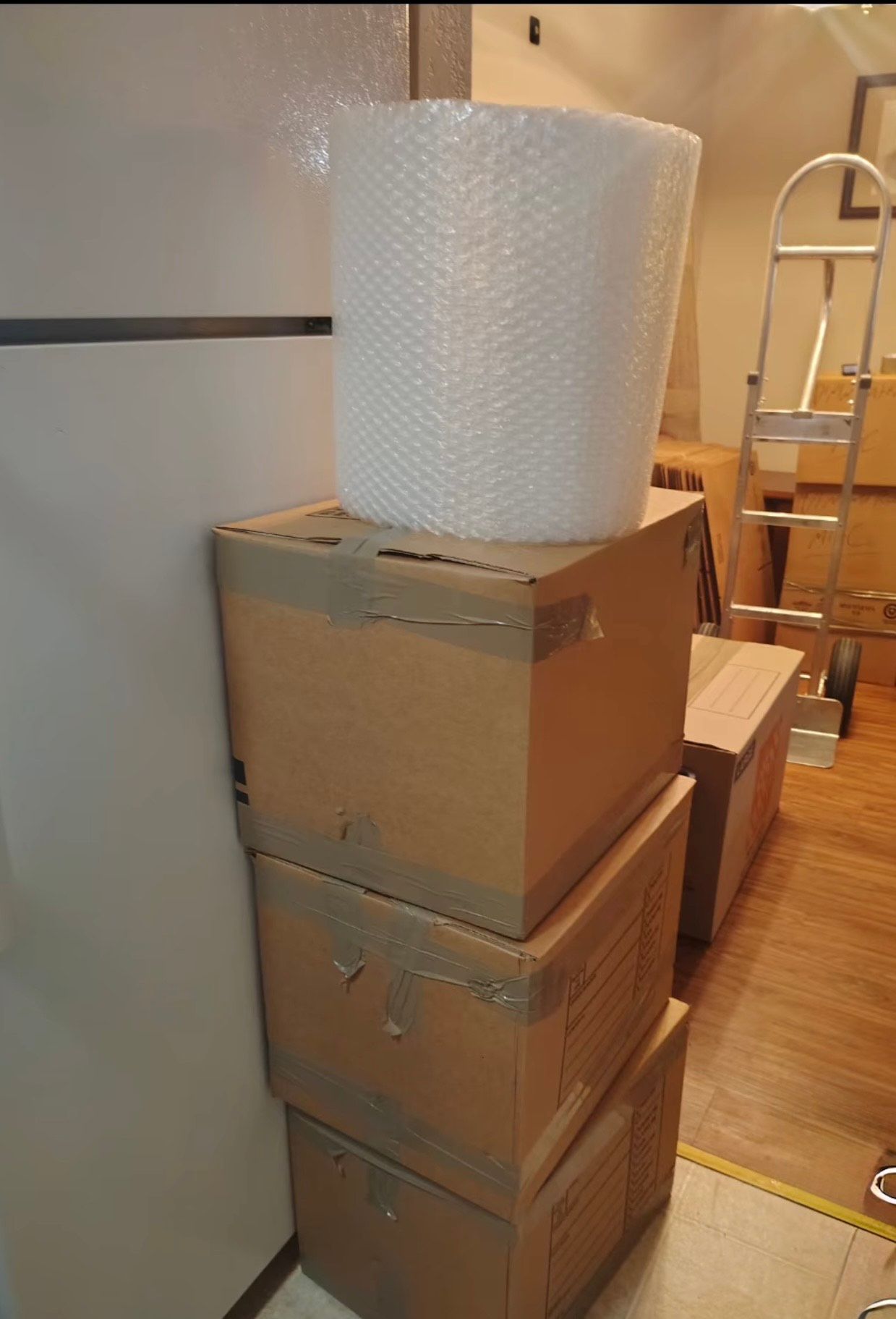 Stack of moving boxes with bubble wrap roll, near a white refrigerator and a hand truck.