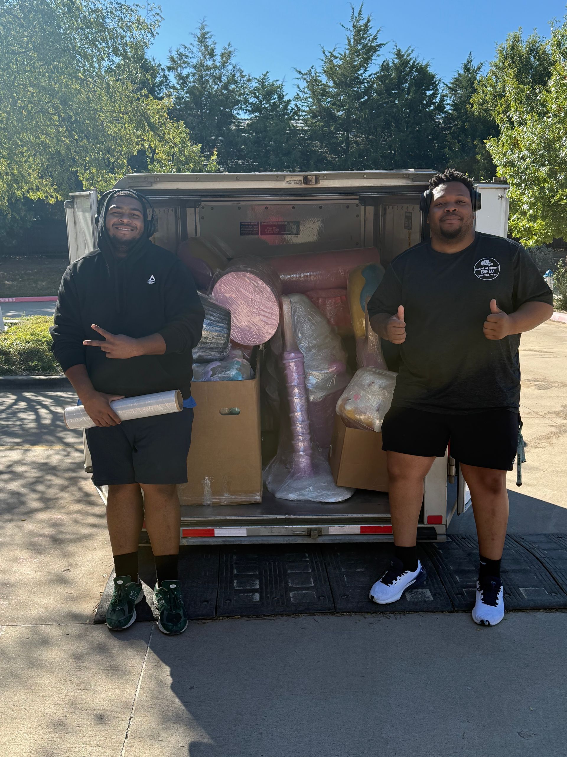 Two men stand in front of a truck loaded with boxes and items. One gives a peace sign, the other a thumbs up.
