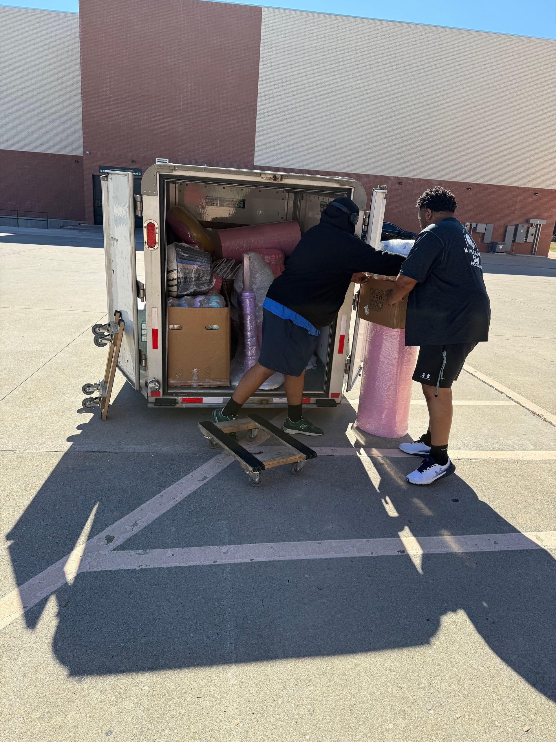 Two people unloading boxes from a white cargo trailer in a parking lot.