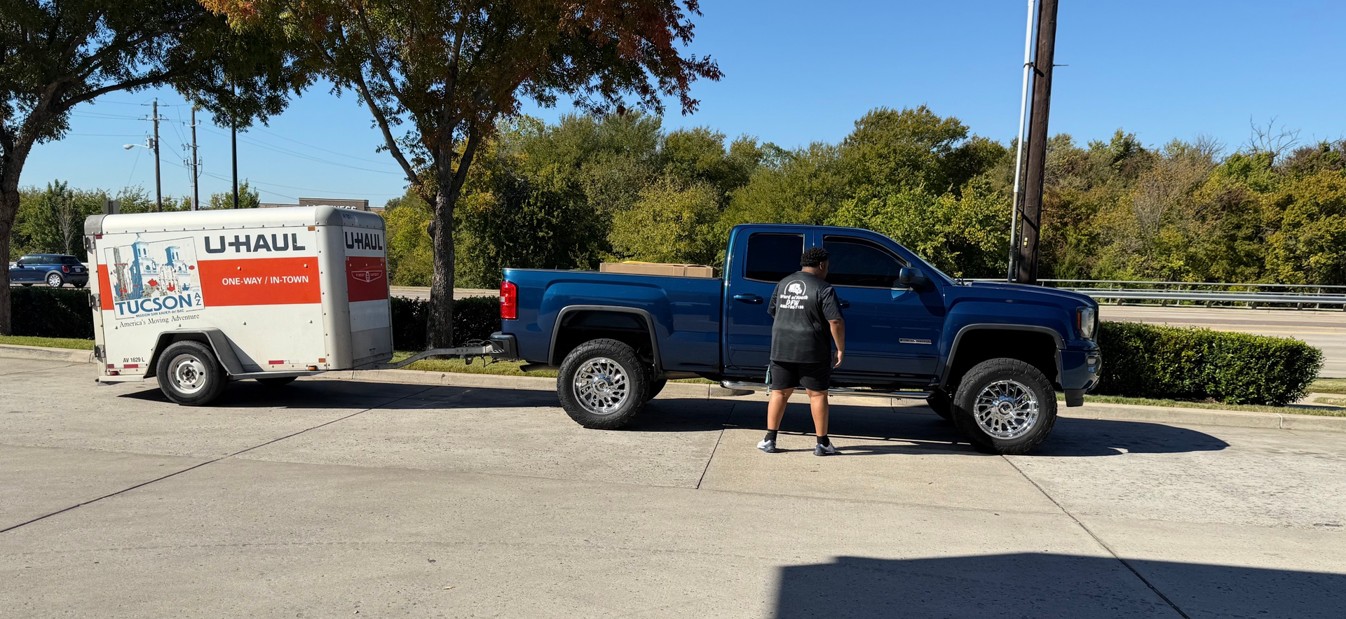 A blue pickup truck is hitched to a U-Haul trailer. A person stands near the truck.