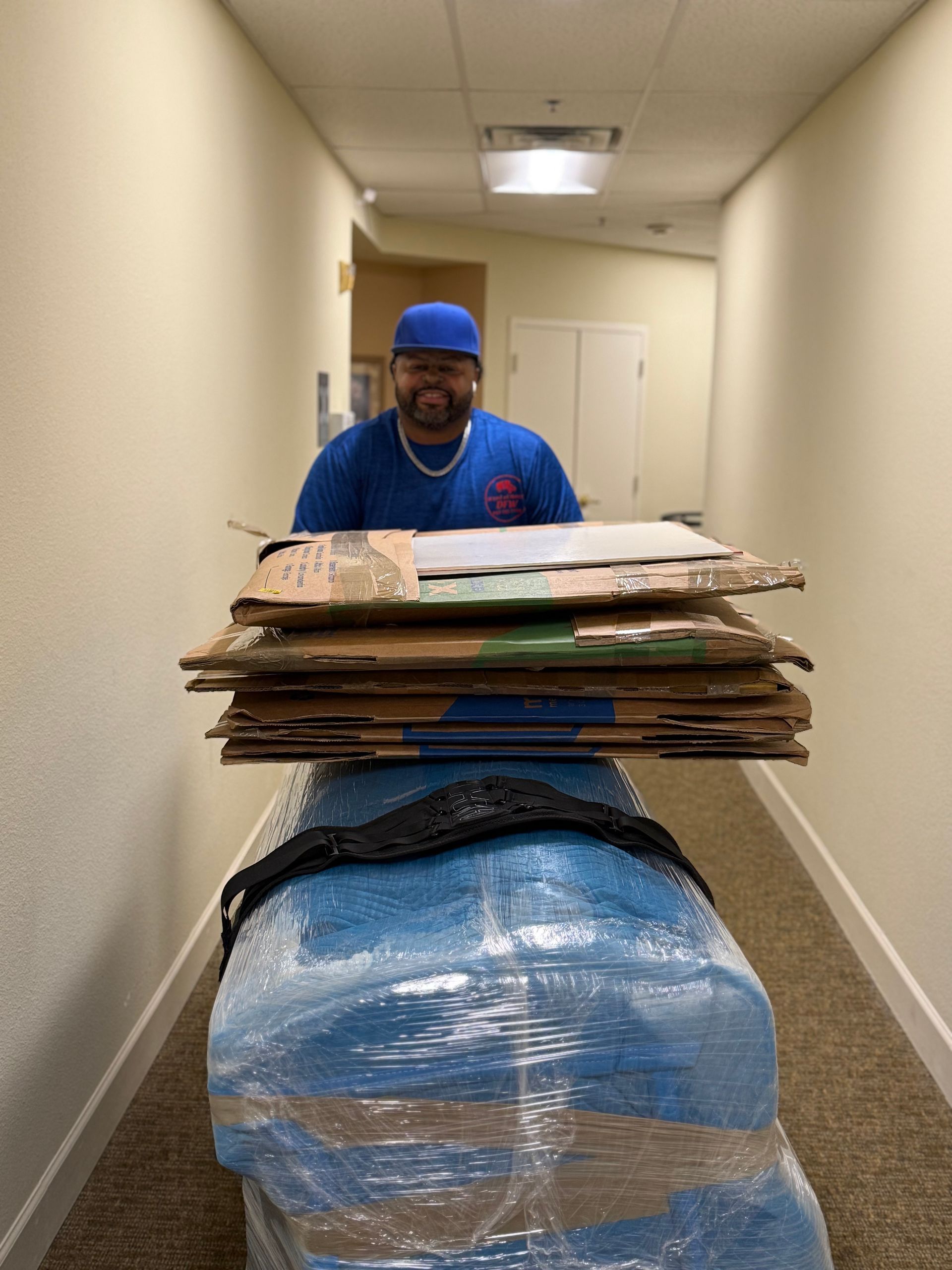 Man smiles, pushing wrapped furniture and cardboard down a hallway with beige walls and a low ceiling.