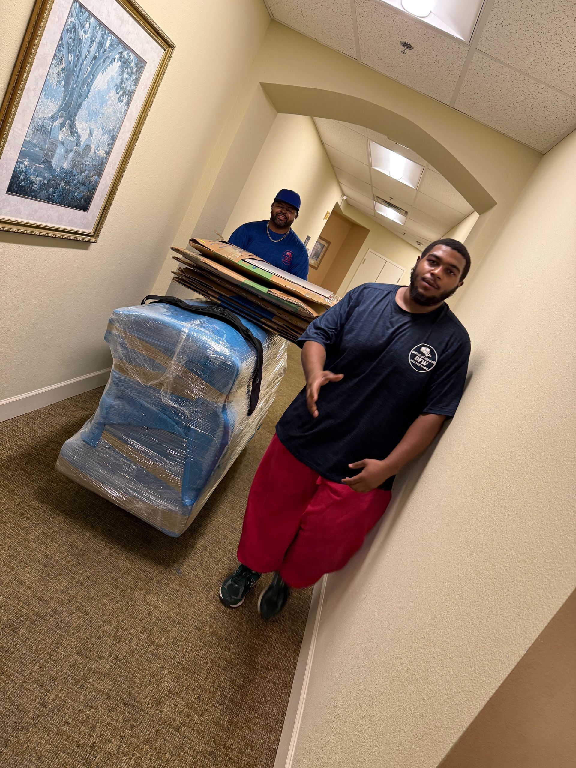Two movers transporting wrapped furniture down a hallway. One gestures, the other pushes the cart.