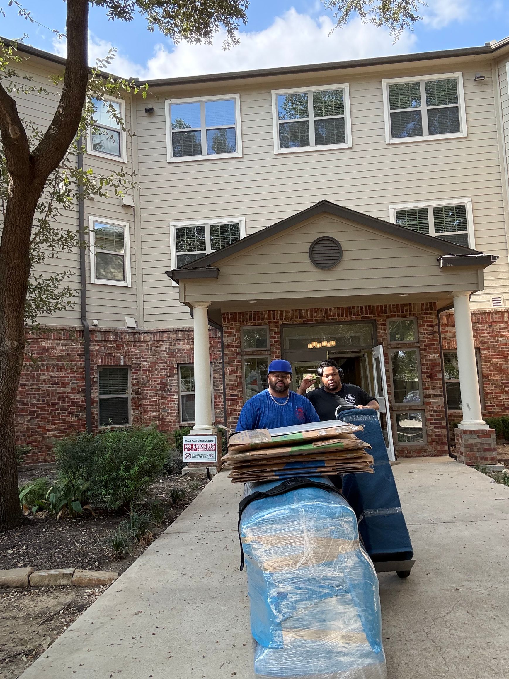 Two movers with wrapped items and a hand truck exiting an apartment building entrance.